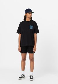 Černé bavlněné tričko s modrým grafickým logem na kapse, doplněné o černé šortky a černé tenisky. Model má na sobě tmavě modrou kšiltovku.