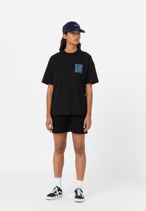T-shirt in cotone nero con logo grafico azzurro sulla tasca, abbinato a pantaloni corti neri e sneakers nere. Il modello indossa un cappellino blu scuro.