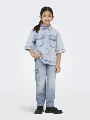 Enfant aux longs cheveux foncés debout en souriant, portant une chemise en denim clair à boutons, un jean cargo et des chaussures noires à lacets sur fond blanc.