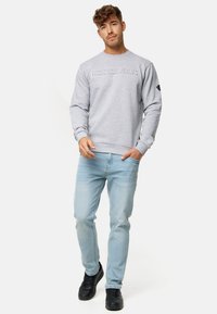Sudadera gris con texto en relieve "INDICODE JEANS", cuello redondo, puños y dobladillo acanalados. Combinada con jeans de color azul claro y zapatillas negras.
