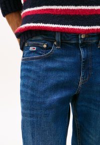 Oseba, ki nosi temno modre kavbojke iz denima z majhno zastavico na žepu in pleten pulover temno modre barve z rdečimi, belimi in modrimi črtami.