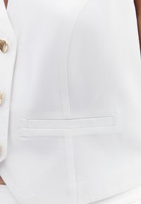 Weißer ärmelloser Blazer aus glattem Stoff; mit goldenen Knöpfen und einer Seitentasche. Der taillierte Schnitt betont die strukturierte Silhouette.