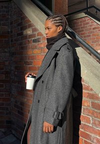 Szary płaszcz w jodełkę z szerokim kołnierzem, rękawami z paskiem i dużymi guzikami. Trzymając biały kubek na kawę przy ceglanej ścianie.