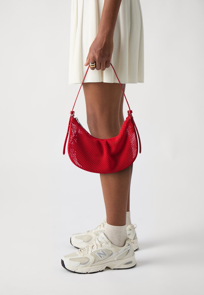Sac à main en filet rouge de forme incurvée, avec une fermeture éclair et une longue bandoulière. Porté avec une robe crème et des baskets blanches.