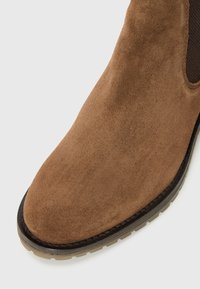 Bota de ante marrón hasta el tobillo con puntera redondeada, panel lateral elástico y suela de goma texturizada. Presenta un borde de contraste negro a lo largo del borde.