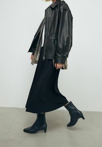 Veste en cuir noire à fermeture éclair à l'avant, portée sur une chemise rayée et une longue jupe noire. Avec des bottines noires élégantes et pointues.