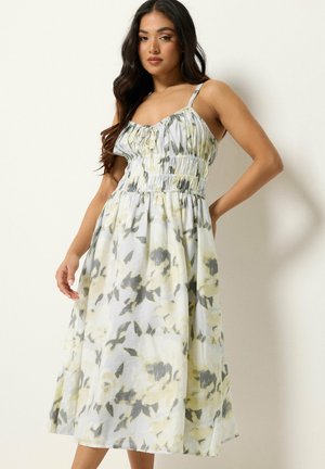 Vrouw draagt een mouwloze, geplooide, bloemige midi-jurk in zacht geel, grijs en wit, staand tegen een effen lichte achtergrond.