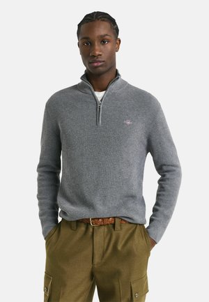 Jong persoon in een grijze sweater met rits tot kwart, een bruine gevlochten riem en olijfgroene cargobroek met handen in de zakken, neutrale achtergrond.