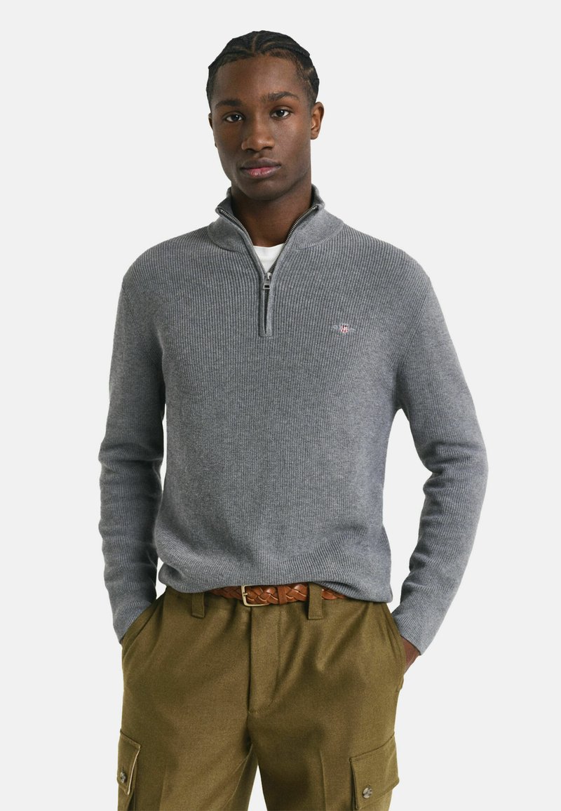 Junge Person in einem grauen Pullover mit Viertelreißverschluss, braunem geflochtenem Gürtel und olivgrünen Cargo-Hosen mit den Händen in den Taschen, neutraler Hintergrund.