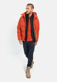 Rød puffjakke med sort inderside, med lynlåslommer og hætte, båret over en orange skjorte og marineblå bukser; tan vandrestøvler.