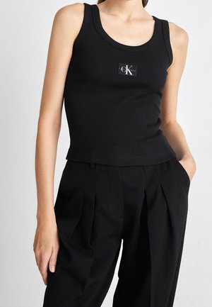 Kvinde iført en sort ribbet tanktop med Calvin Klein-logo og højttaljede sorte plisserede bukser, den ene hånd i lommen.