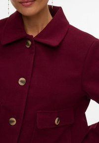 Frau trägt einen tief bordeauxroten Mantel mit weitem Kragen, großen braunen Knöpfen und einer mit Knöpfen versehenen Klappentasche auf der Brust.