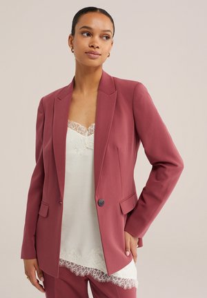 Femme portant un blazer rose poudré sur un caraco blanc bordé de dentelle, avec un pantalon rose poudré assorti, posant avec une main sur la hanche.
