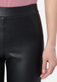 Black faux leather leggings with a high waist, smooth texture, and slim fit. Hand lightly grasping the fabric at the side seam.