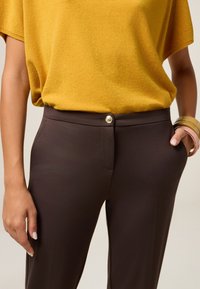 Pantalons marron ajustés avec une texture lisse, dotés d'une fermeture à bouton à l'avant et de poches latérales. Associés à un haut jaune à manches courtes.