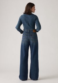 Salopette en denim bleu foncé avec un corsage ajusté et un pantalon à jambes larges. Présente un empiècement brodé et une étiquette en cuir sur la taille arrière.