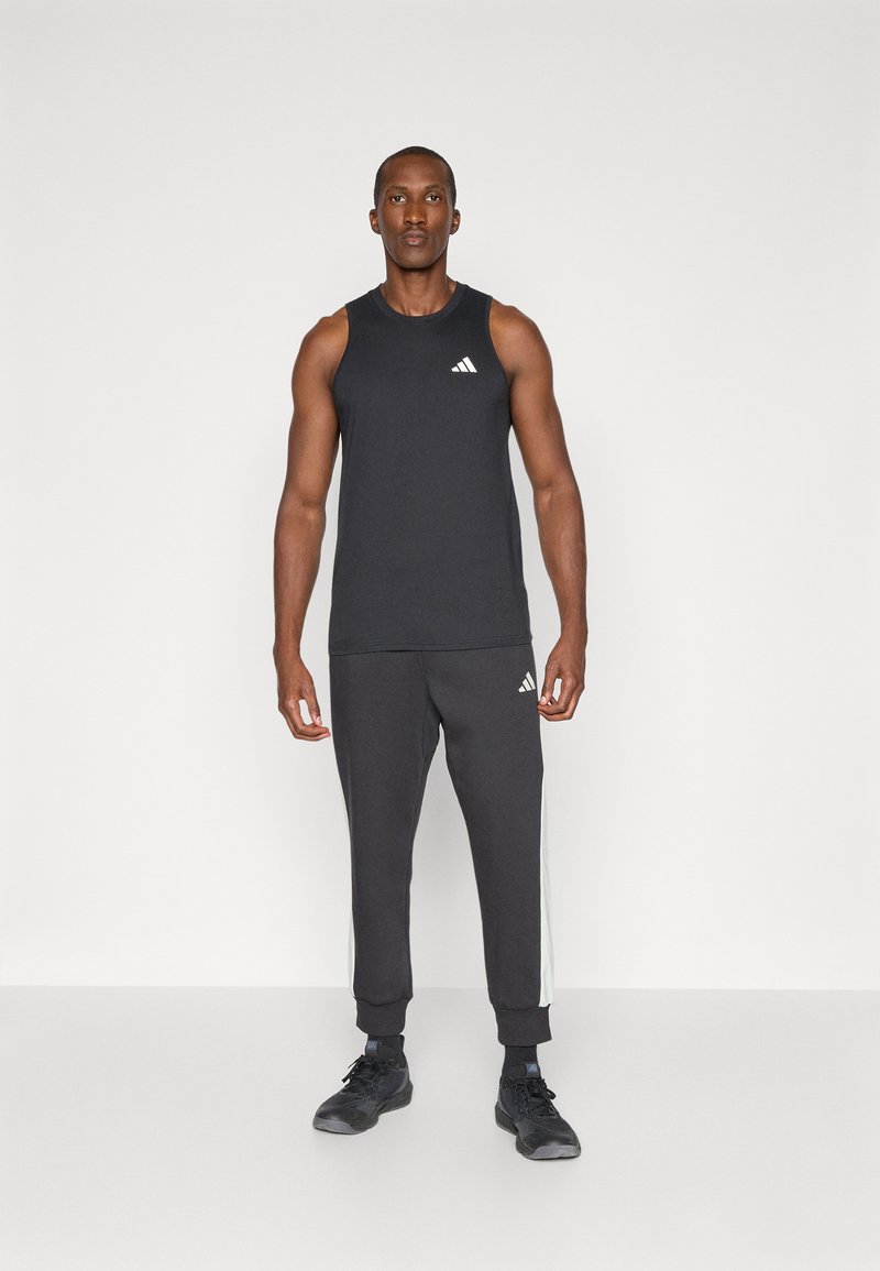 Czarny bezrękawnik sportowy z białym logo, zestawiony z ciemnoszarymi dresami z białymi paskami po bokach i czarnymi butami sportowymi.