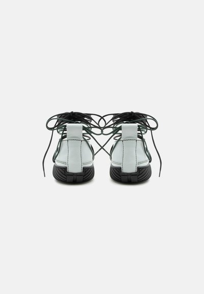 Άσπρα δερμάτινα σανδάλια με μαύρες σόλες, φαίνονται από πίσω, με μαύρα κορδόνια δεμένα πάνω από τις φτέρνες σε μονόχρωμο φόντο.