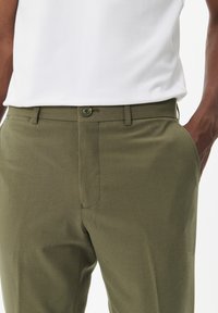 Pantalones verde oliva con una textura suave, que cuentan con cierre de botón, bolsillos laterales y un corte ajustado. Llevados con una camiseta blanca.