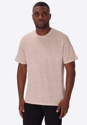 Uomo con capelli corti e barba che indossa una maglietta beige tinta unita a maniche corte e pantaloni neri, in piedi davanti a uno sfondo bianco.