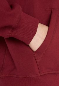 Burgundy hoodie with a soft texture, featuring a kangaroo pocket and ribbed cuffs. Seam detailing visible along collar and pocket edges.