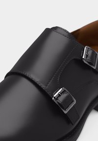 Chaussure habillée à boucle de moine en cuir noir avec deux boucles métalliques argentées et coutures détaillées sur fond blanc.