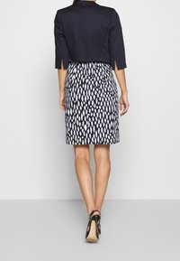 Donkerblauwe cropped jas met driekwart mouwen, gedragen over een knie-length zwart-wit gestreepte jurk met een diamanten patroon.