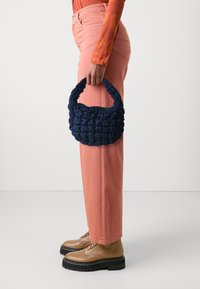 Une personne portant un pantalon corail et des chaussures beiges tient un sac à main matelassé bleu marine avec une anse tressée à son côté.