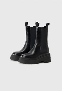 Bottines noires à semelle épaisse et panneaux latéraux élastiques, avec un détail de marque. Positionnées sur un fond uni.