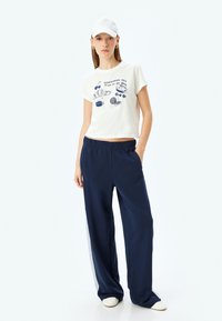 Geknipt wit T-shirt met blauwe grafische tekst en voedselillustraties, gecombineerd met marineblauwe wijde broek met een lichtgrijze zijscheiding.