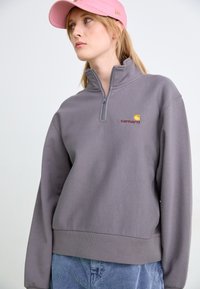 Sudadera gris con media cremallera, de corte relajado, puños acanalados y un pequeño logo bordado. Combinada con pantalones texturizados azul claro y una gorra rosa.