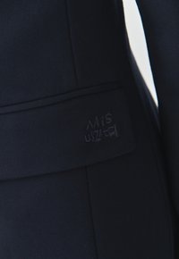 Gros plan sur un tissu foncé avec un patch rectangulaire cousu affichant un texte stylisé partiellement visible en fil foncé.