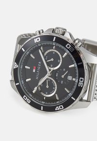 Silberner Chronograph mit schwarzem Zifferblatt, silbernen Stundenmarkierungen, drei zusätzlichen Zifferblättern und einem strukturierten Netzarmband. Verfügt über eine drehbare Lünette.