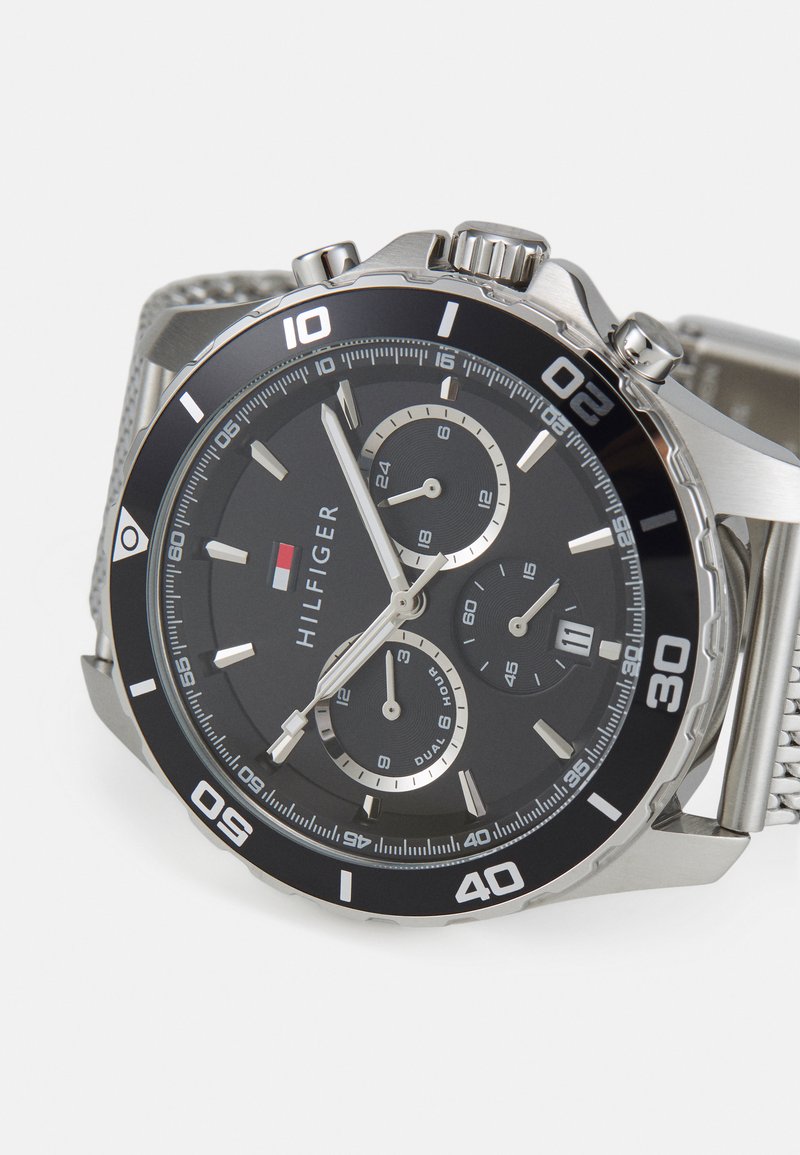 Reloj cronógrafo plateado con un dial negro, marcadores de hora plateados, tres subdiales y una correa de malla texturizada. Incluye un bisel giratorio.