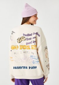 Sudadera de gran tamaño color crema con estampados gráficos coloridos, incluyendo texto, dados y llamas en la parte de atrás. Puños acanalados y tejido texturizado.