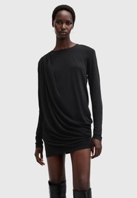 Abito lungo a maniche lunghe nero drappeggiato con una texture liscia, caratterizzato da un collo asimmetrico e dettagli arricciati, abbinato a stivali al ginocchio.