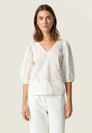 Vit blus med V-ringning och puffärmar, med broderi och utskärningar längs fållen. Tillverkad av lättviktsmaterial.