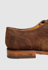 Braune Wildlederschuhe mit glatter Textur, dekorativen Perforationen und einem gestapelten Holzabsatz, die ein abgerundetes Zehendesign zeigen.