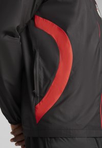Veste noire avec des accents rouges, présentant une texture lisse et un design incurvé. Comprend une poche zippée sur le côté pour le rangement.
