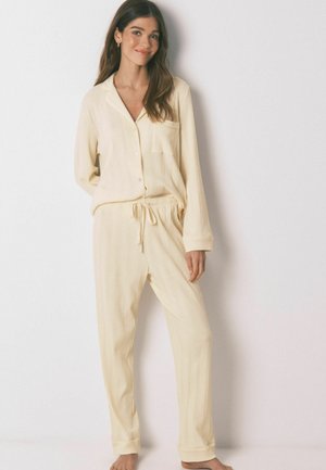 Kvinde stående barfodet iført cremefarvet ribbet knap-ned top med lange ærmer og matchende bukser med snøre mod en ensfarvet baggrund.