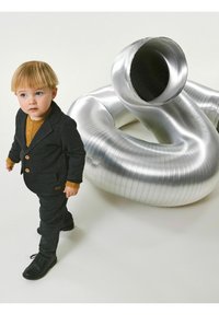 Un bambino in un completo grigio scuro con bottoni marroni, abbinato a un maglione color senape, si trova vicino a un tubo metallico argento arrotolato.