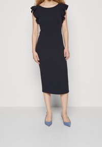 Laivansininen midi-mekko, jossa on istuva siluetti, röyhelökoristeiset hihansuut ja sileä pinta. Yhdistetty kärkitossuihin, jotka ovat myös siniset.