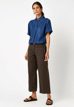 Woman standing wearing a short-sleeve blue collared shirt, brown wide-leg pants, and brown sandals against a plain white background.