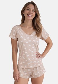 Helle beige Schlafanzug-Set mit V-Ausschnitt, das ein Herzmuster in Weiß aufweist. Weicher Stoff, kurze Ärmel und passende Shorts mit elastischem Bund.