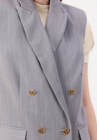 Harmaa pinstripe-vesti, jossa on kaksirivinen malli, kultasävyiset napit, terävät läpät ja jämäkkä siluetti. Teksturoitu kankaan pinta.