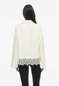 Kremhvit bluse med lange ermer, satengtekstur, med en blondekant nederst som en dekorativ detalj. Brukt med svarte leggings.