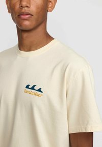 Crème korte mouw t-shirt van katoen, met een blauw golfontwerp en de tekst "Revolution" in het geel. Zachte textuur, ronde halslijn.