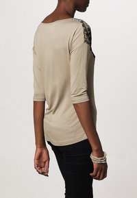 Beige long-sleeve top with black lace detailing on shoulders, round neckline, and a relaxed fit. Fabric appears soft with subtle texture.