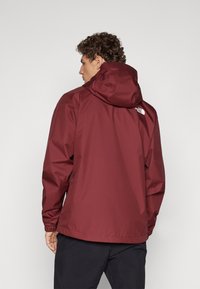 Chaqueta impermeable color burdeos con capucha, con puños elásticos y un logo en la parte posterior. Textura suave y corte relajado.