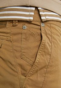 Detailní záběr na kapsu béžových kalhot s kovovými nýty a švy, nošených s pruhovanou béžovo-bílou košilí zasunutou dovnitř.